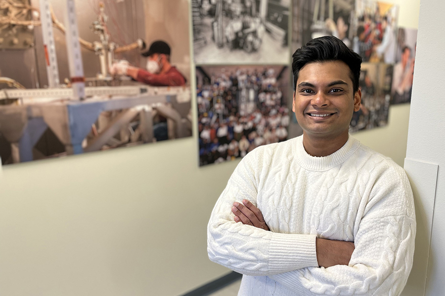 A smiling researcher in a white sweater stands with arms crossed in front of a wall displaying laboratory and research photographs.