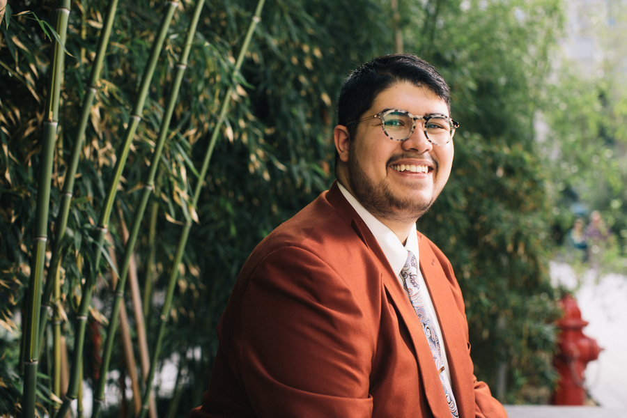A man in a rust-colored blazer and glasses smiles broadly in front of a grove of bamboo.