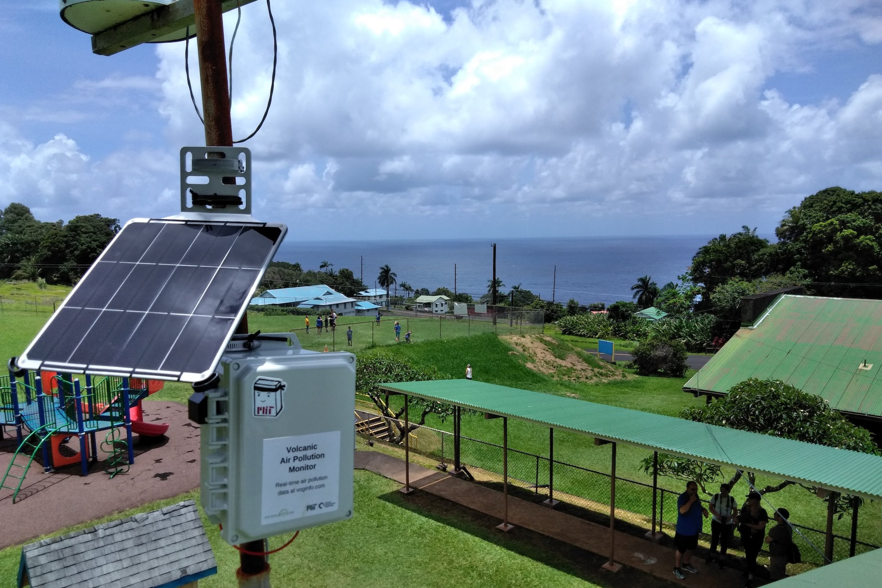 A solar-powered volcanic air pollution monitor mounted on a pole at a Hawaiian school, with the ocean and green landscape visible in the background.