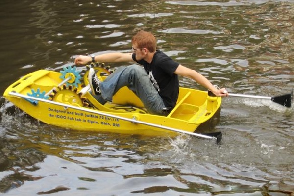 A person wearing sunglasses rows a small, bright yellow boat with visible gears, creating splashes on a calm body of water. 