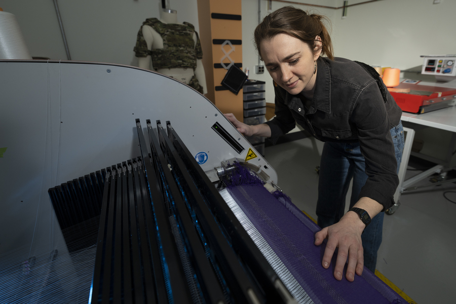 A researcher leans over an industrial knitting machine producing purple fabric, with a textile vest and lab materials visible in the background.