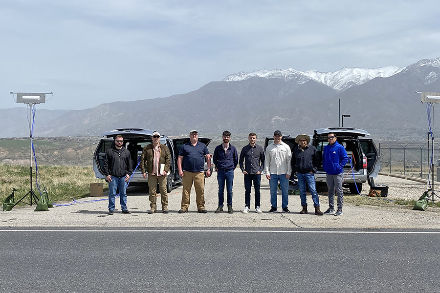 A research team of eight people stands in a line on a roadside with two SUVs and mounted antenna arrays behind them, with snow-capped mountains visible in the background during a 5G field testing campaign