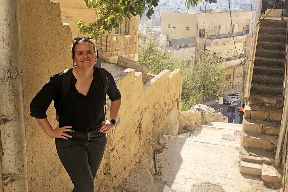 A woman in a black shirt smiles while leaning against a sun-lit stone staircase in an old Middle Eastern city neighborhood with limestone buildings and rooftops in the background.