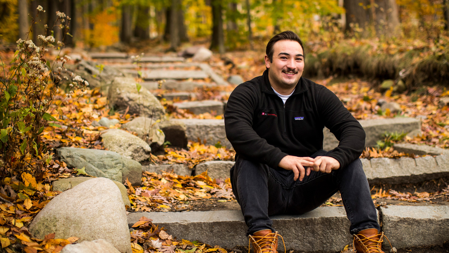 A smiling young man in a black fleece jacket sits on stone steps surrounded by fallen autumn leaves and boulders in a wooded campus setting