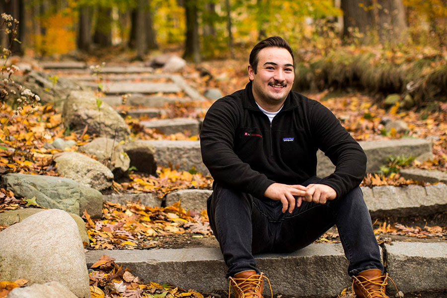 A smiling young man in a black fleece jacket sits on stone steps surrounded by fallen autumn leaves and boulders in a wooded campus setting