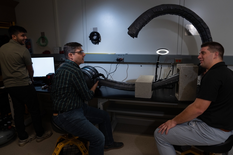 Three researchers in a lab gather around a flexible robotic arm arching overhead, with a computer monitor and lab equipment visible in the background.