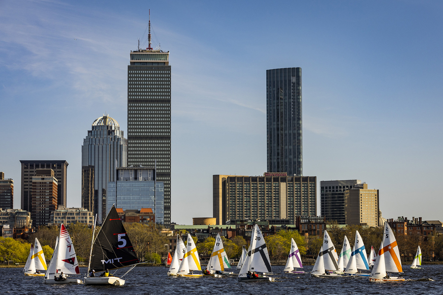 A fleet of MIT sailing team dinghies race on the Charles River with the Boston skyline, including the Prudential Tower, in the background.