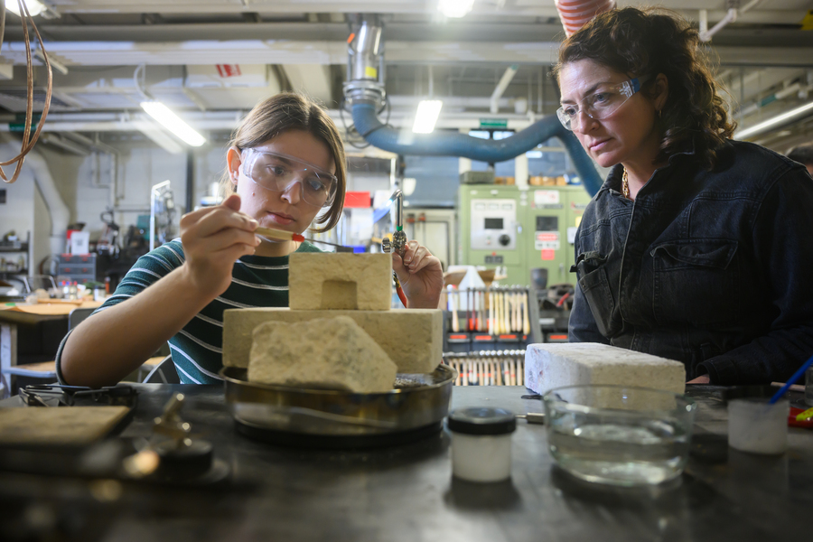 Two MIT researchers wearing safety glasses work together at a lab bench, with one carefully probing stacked sandstone rock samples while the other observes closely, surrounded by lab equipment and tools.