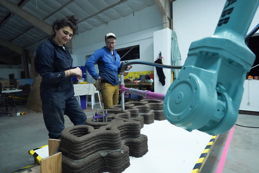 Two researchers observe a large industrial robotic arm extruding layered mud coils to 3D-print a textured architectural structure inside a warehouse fabrication studio.
