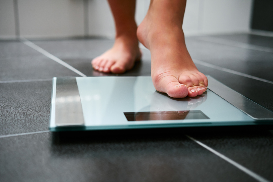 Close-up of bare feet stepping onto a glass bathroom scale on a dark tile floor.