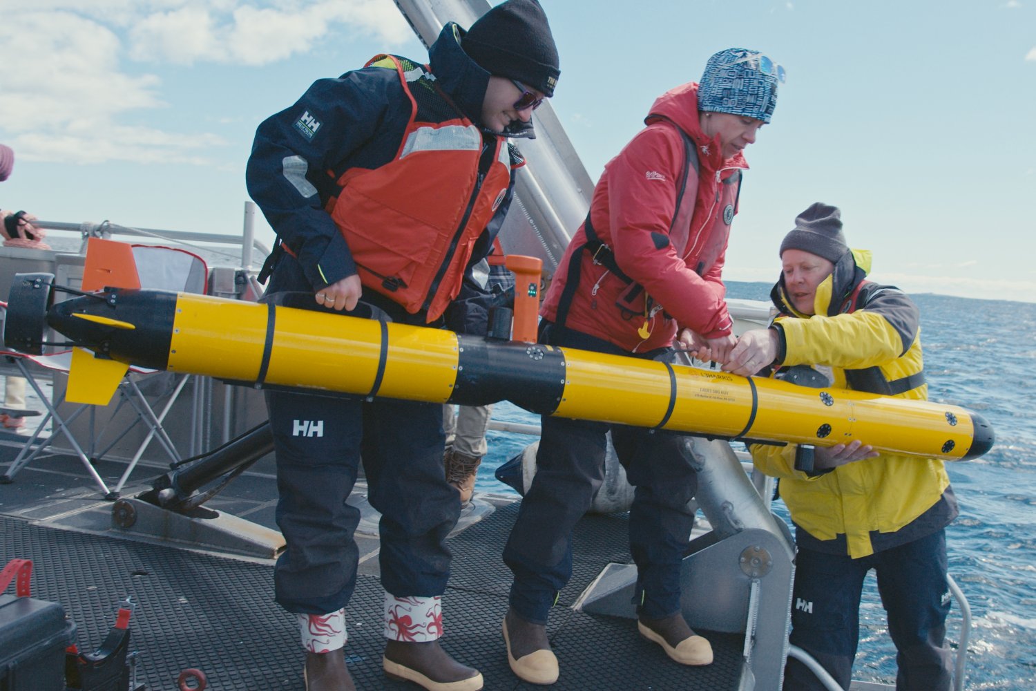 Three researchers in cold-weather gear and life vests prepare to deploy a yellow torpedo-shaped autonomous underwater vehicle from the deck of a research vessel at sea