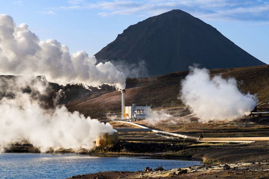 Geothermal power facility releasing steam near a lake, with pipes and a mountain in the background.