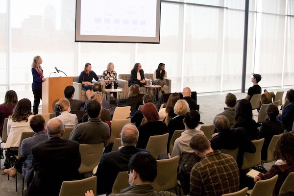 Several panelists are in the front of a large room while rows of people in chairs sit facing them.