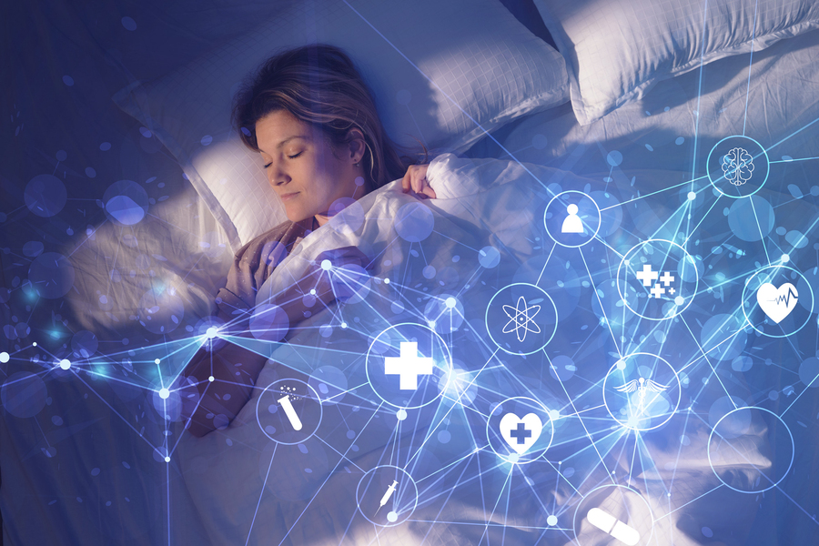 A woman sleeps peacefully in a dimly lit bedroom while a blue glowing network of connected medical icons including a heart monitor, brain, and cross symbols floats above her, representing ingestible smart health monitoring during sleep.