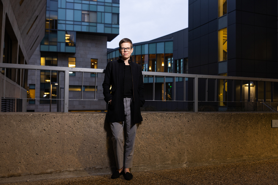 A person in a black coat and glasses stands with hands in pockets against a concrete wall at dusk, flanked by illuminated modern glass academic buildings