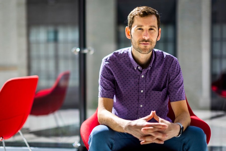 A man in a purple shirt sits forward in a red chair in a bright, modern interior space, looking directly at the camera.