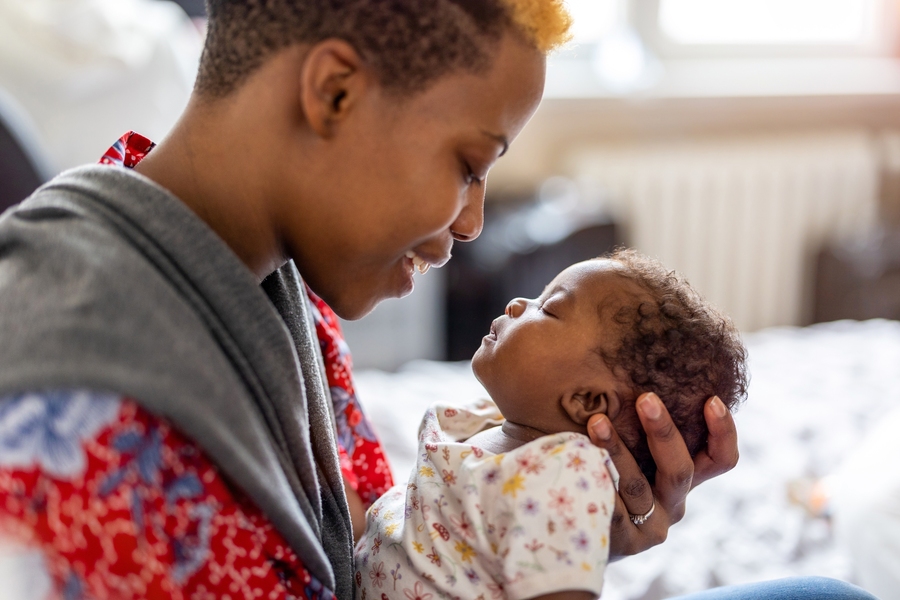A parent smiles while cradling a sleeping newborn face-to-face in a softly lit home setting.