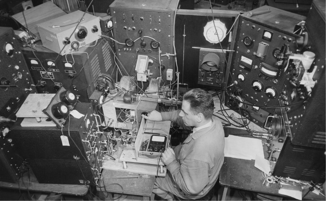 Black-and-white photo of a man operating a complex array of vintage scientific equipment and electronics in a lab setting.