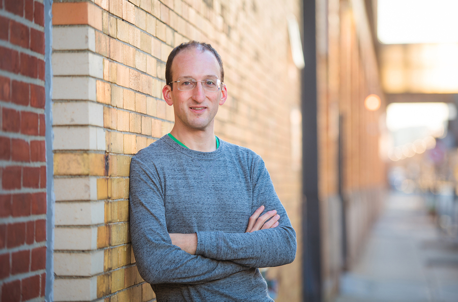 A man with glasses and a gray sweater leans against a brick wall outdoors, arms crossed, smiling at the camera.