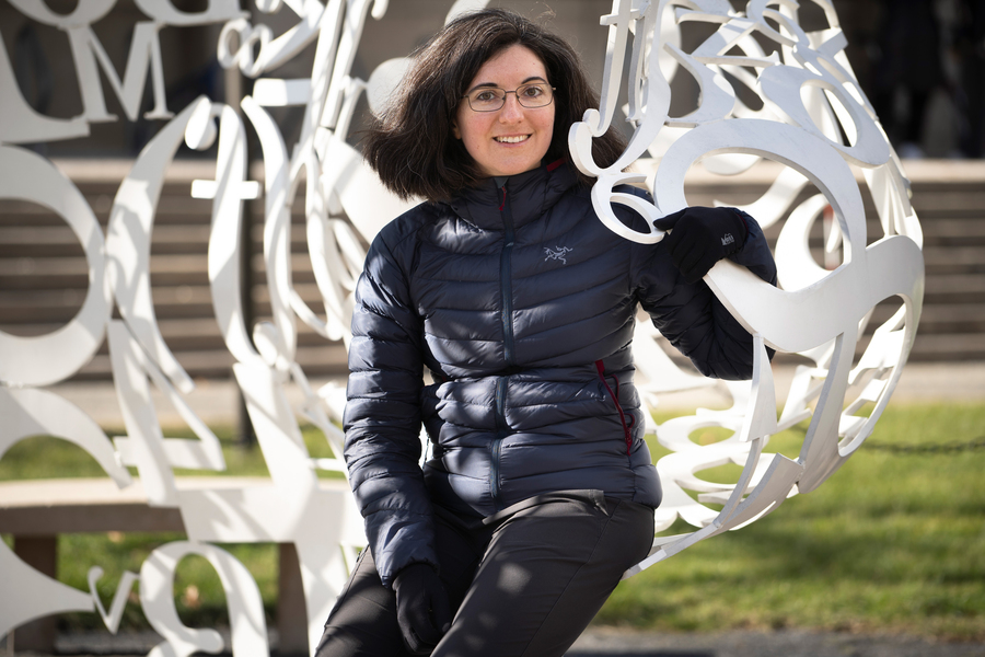 A woman in a navy puffer jacket leans against a large white outdoor sculpture made of interlocking letter forms on a sunny campus plaza.