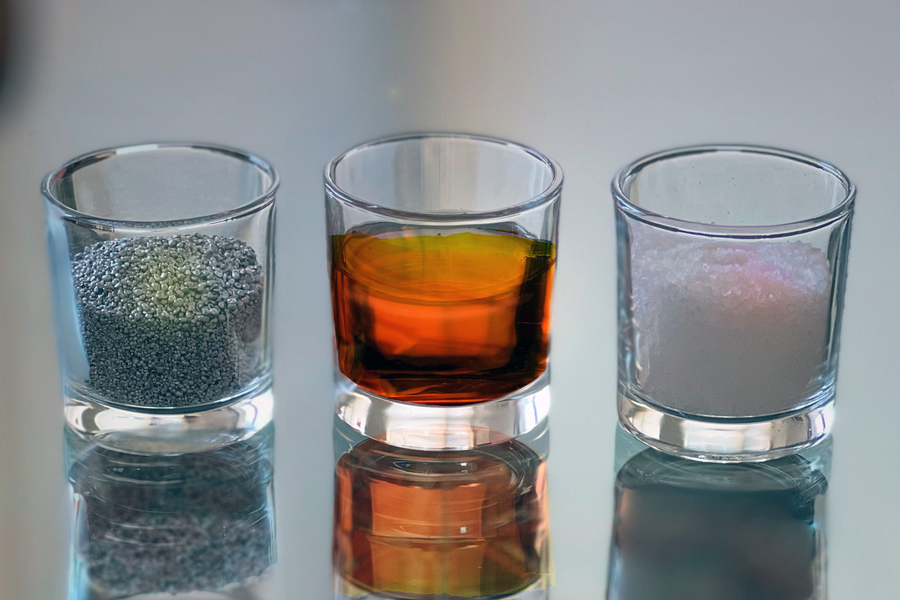 Three small glass tumblers on a reflective surface hold gray granules, an amber liquid, and pale pink crystals, representing battery material components.