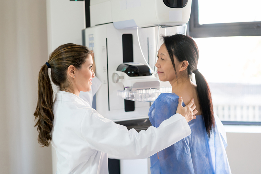A radiologist in a white coat assists a patient in a blue gown positioning for a mammogram at a digital imaging machine.