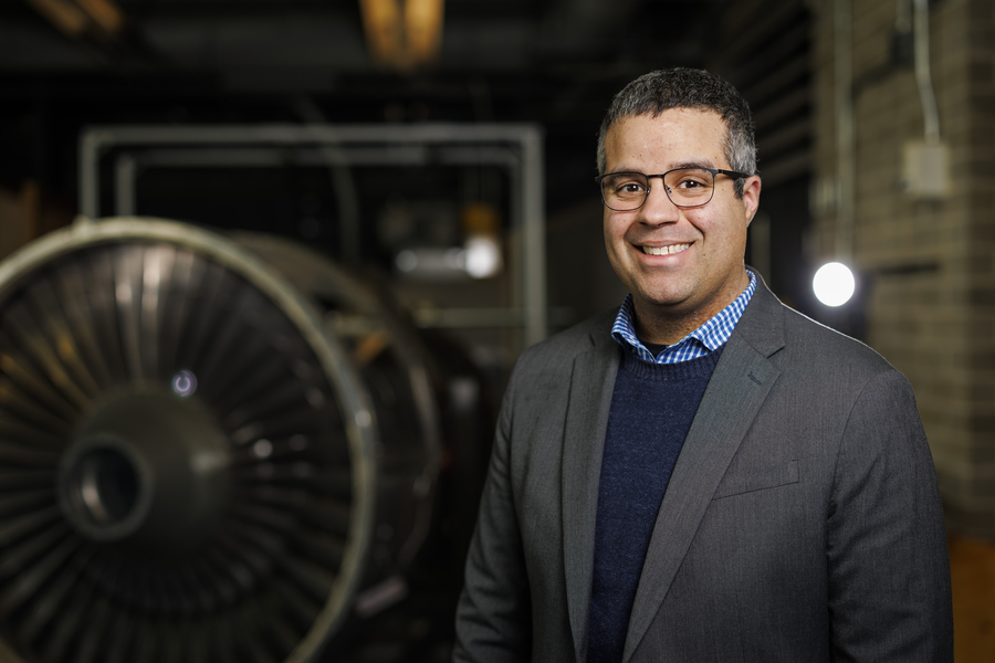 A smiling researcher in a gray blazer stands in front of a large jet engine turbine in an aerospace engineering facility