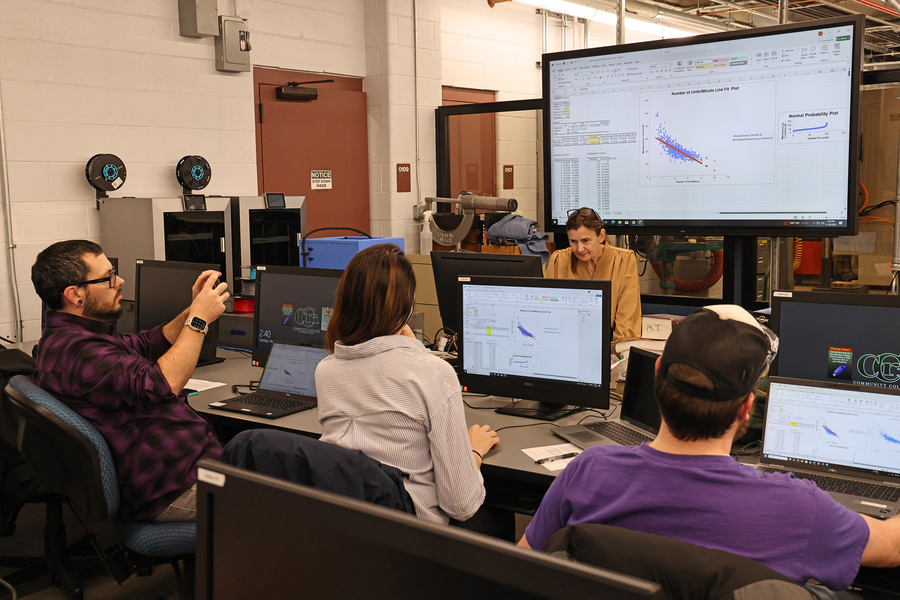Three students and an instructor work at computer workstations in a manufacturing lab, with statistical data charts displayed on a large monitor overhead.