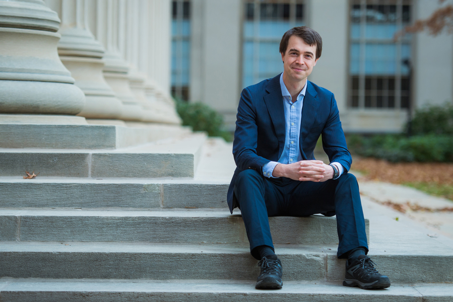 A man in a navy suit sits on the stone steps of a columned academic building with hands clasped, smiling at the camera