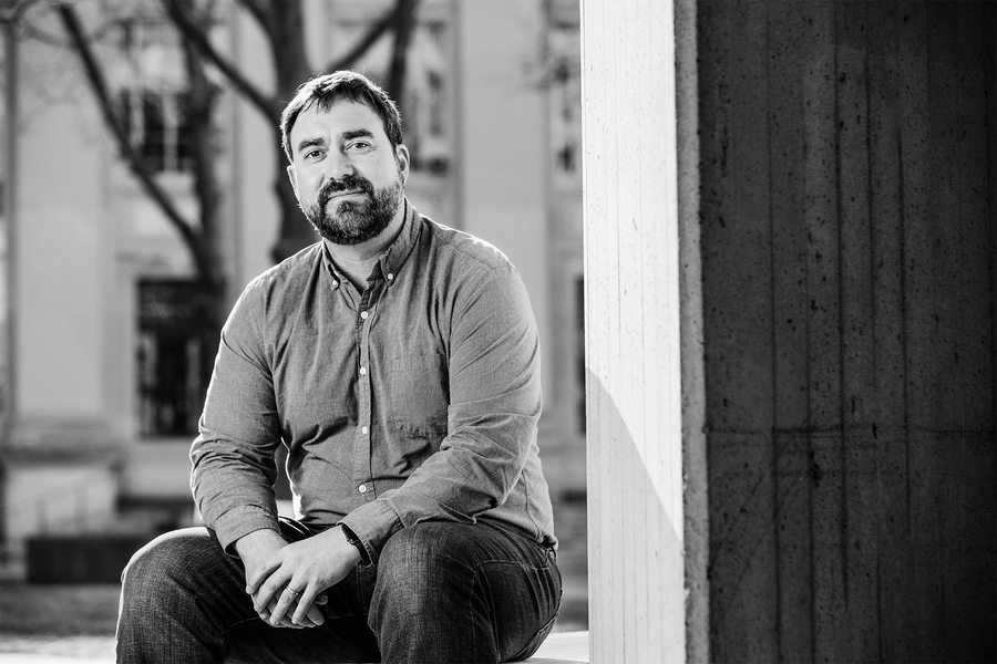Black and white portrait of a bearded man in a button-down shirt sitting against a concrete column outside a campus building.