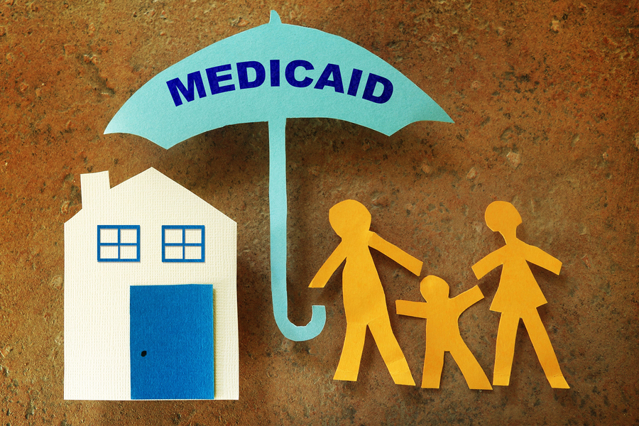 Paper cutout illustration of a family of three standing beneath a blue Medicaid umbrella next to a paper house, set against a textured brown background.