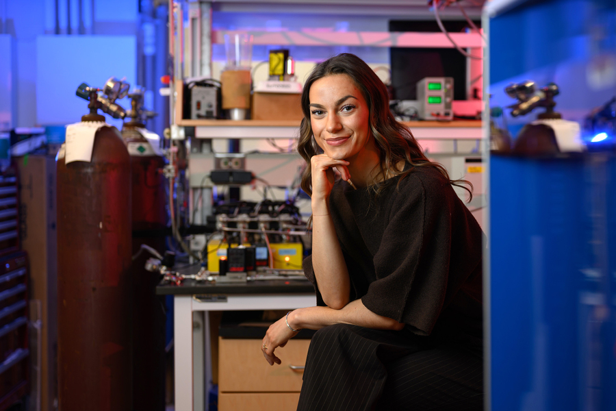 A woman poses in a research lab surrounded by equipment including gas cylinders, electronic instruments, and wiring, lit with dramatic blue and red light.