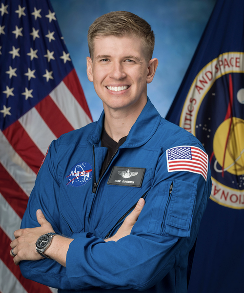 Official NASA portrait of an astronaut candidate in a blue flight suit with arms crossed, standing in front of an American flag and the NASA seal.