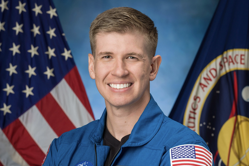 Official NASA portrait of an astronaut candidate in a blue flight suit with arms crossed, standing in front of an American flag and the NASA seal.