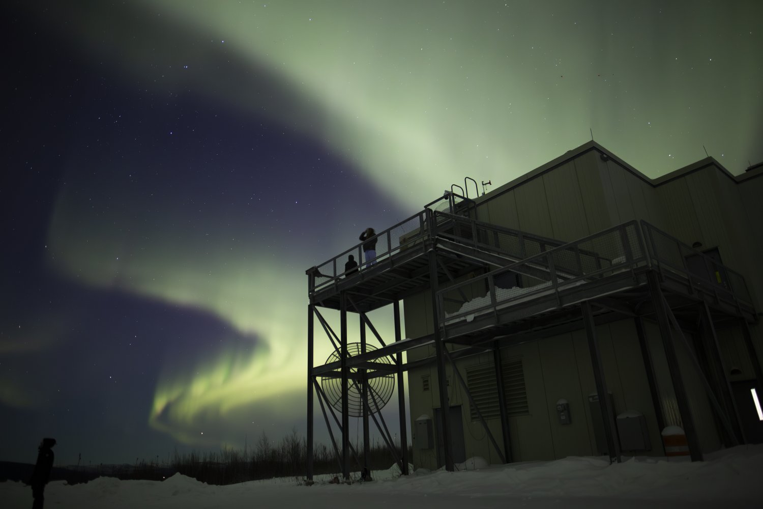 The aurora borealis glows green over a research station as people watch from an elevated platform.