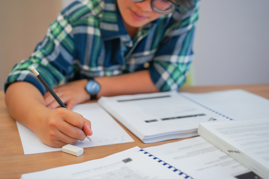 A student wearing glasses and a plaid shirt writes in a notebook at a desk surrounded by open textbooks and coursework.