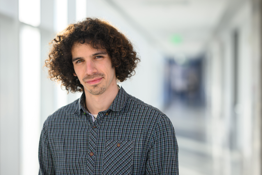 A young MIT researcher with curly dark hair wearing a plaid shirt, photographed in a bright corridor with blurred background.