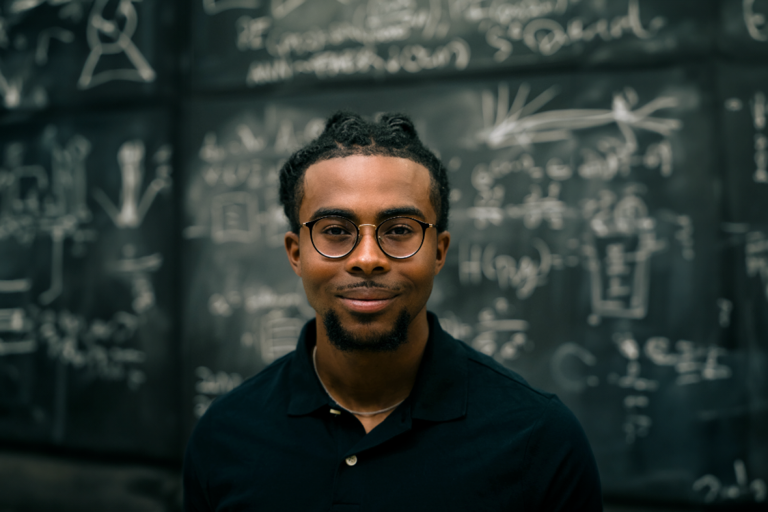 Portrait of a man wearing glasses and a black shirt in front of a chalkboard covered with equations.