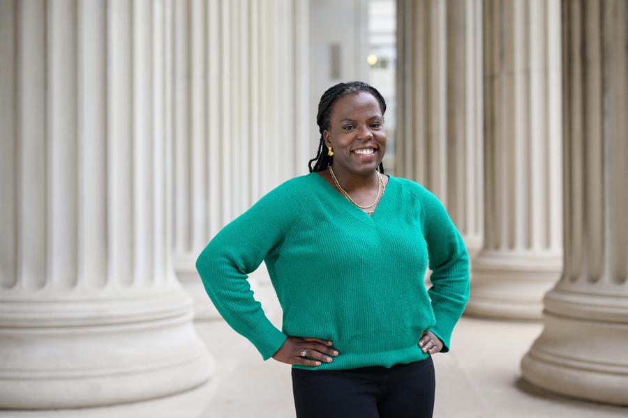 A smiling MIT researcher in a green ribbed sweater stands with hands on hips in front of the stone columns of MIT's Building 10 colonnade.
