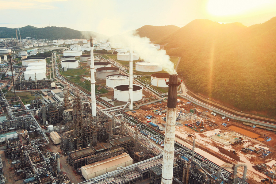 Aerial view at sunset of a large industrial oil refinery complex with smokestacks emitting white steam, storage tanks, and pipeline infrastructure nestled against forested hills