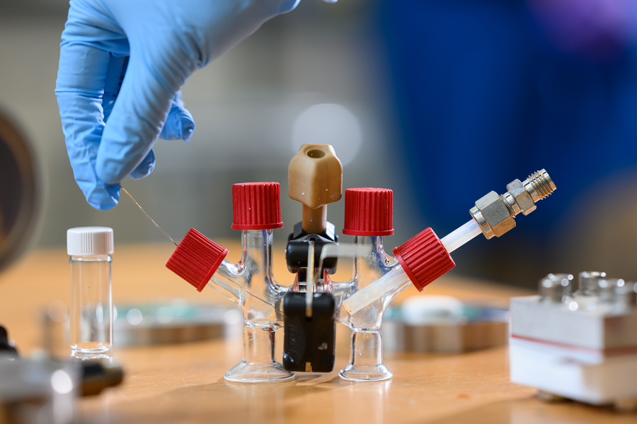 A gloved hand holds a thin wire over a small glass apparatus with red-capped vials and metal fittings on a lab bench.