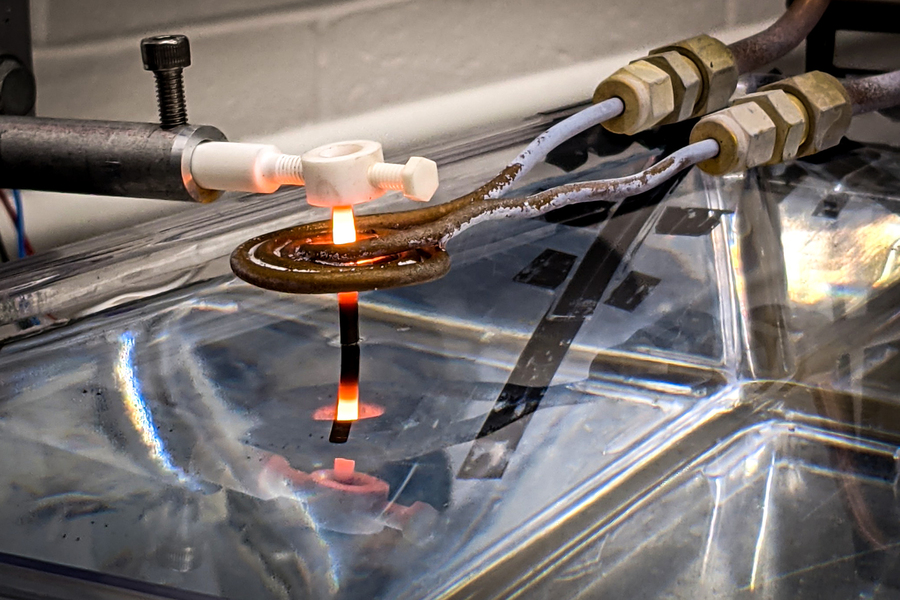 Close-up of a laboratory heat treatment experiment showing a glowing orange-red metal strip submerged beneath the surface of a clear liquid tank with industrial tubing and fittings above.