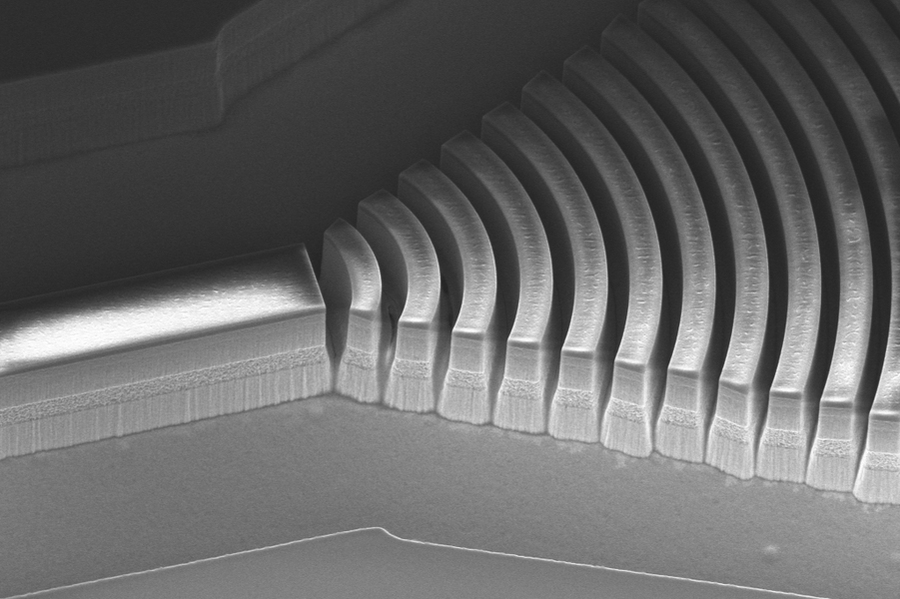 Scanning electron microscope image of a photonic integrated circuit showing tightly spaced curved diffraction grating ridges etched into a semiconductor substrate, captured in high-resolution grayscale.