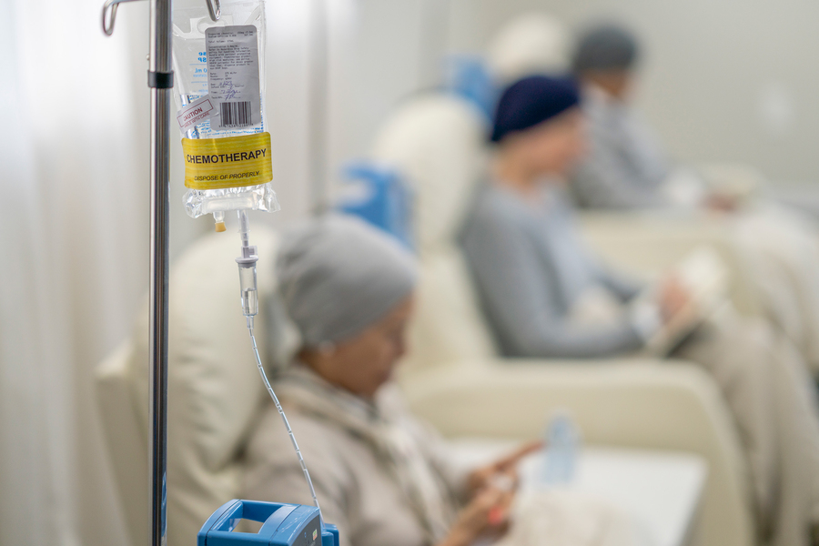 A labeled chemotherapy IV bag hangs in the foreground while patients wearing head coverings sit in treatment chairs in a blurred background.
