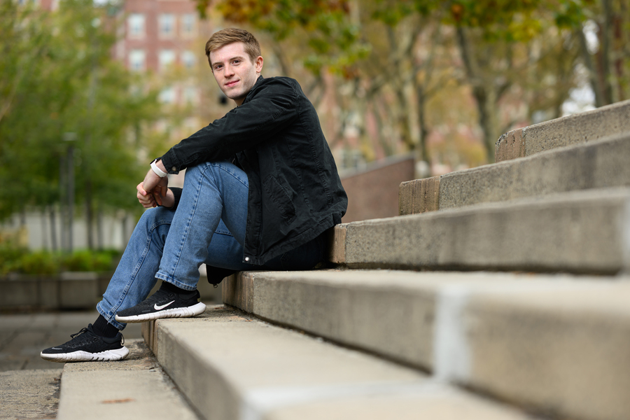 MIT student seated casually on outdoor concrete steps in fall, wearing a black jacket and jeans, looking at the camera.