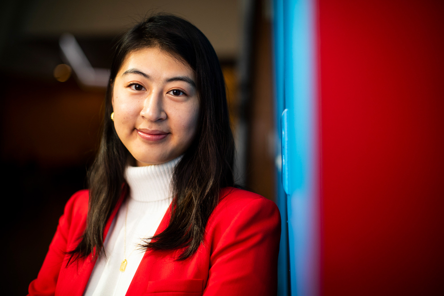 A woman in a red blazer and white turtleneck looks at the camera, with a bold red and blue background behind her.