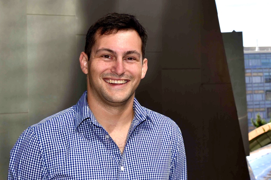 A man in a blue gingham shirt smiles broadly while leaning against a dark angular structure outdoors with a building visible behind him.