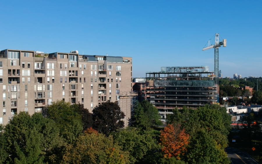 Aerial view of a mid-rise residential building beside a multi-story building under active construction with a tower crane, surrounded by mature trees in early autumn foliage.