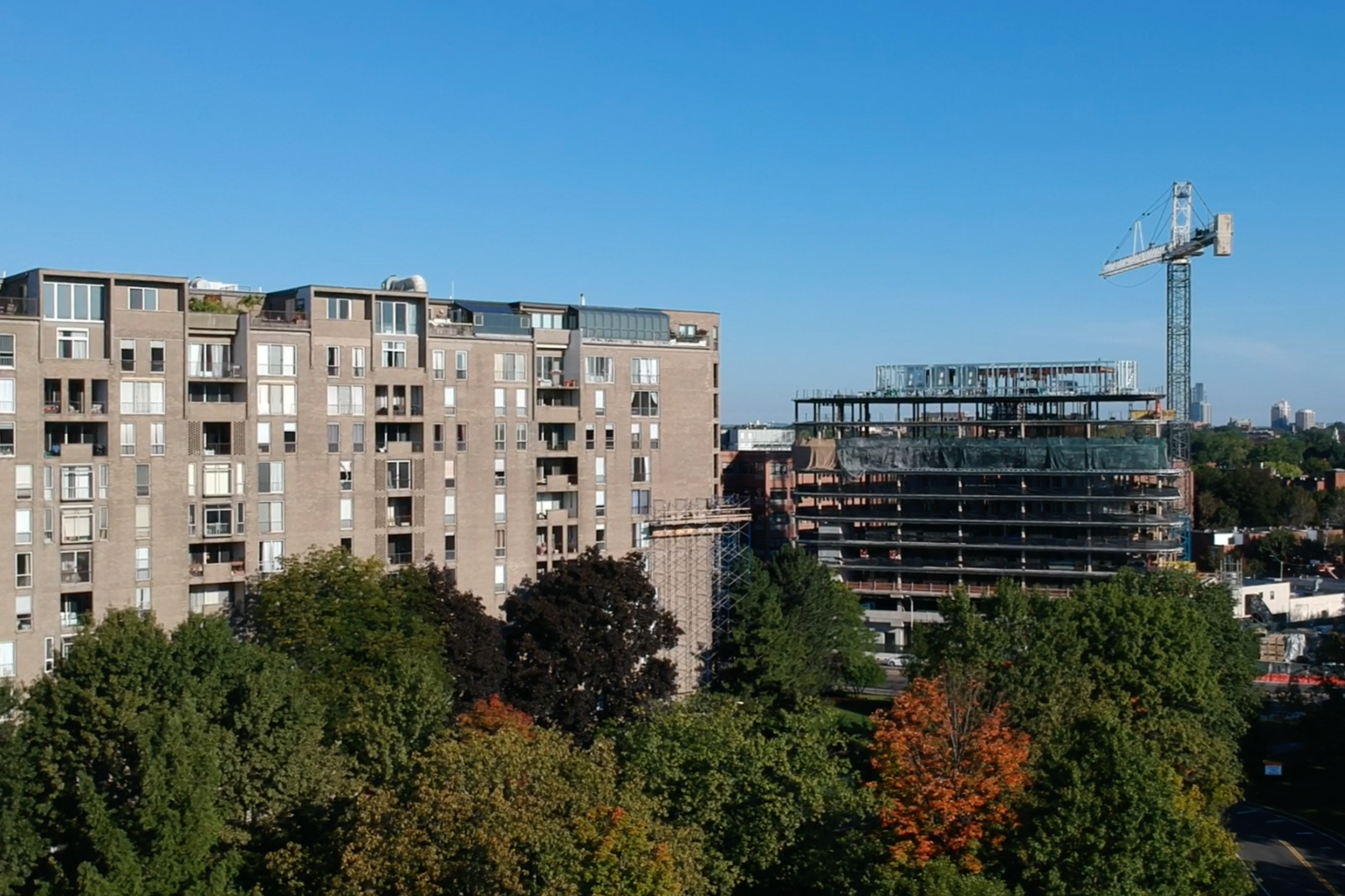 Aerial view of a mid-rise residential building beside a multi-story building under active construction with a tower crane, surrounded by mature trees in early autumn foliage.
