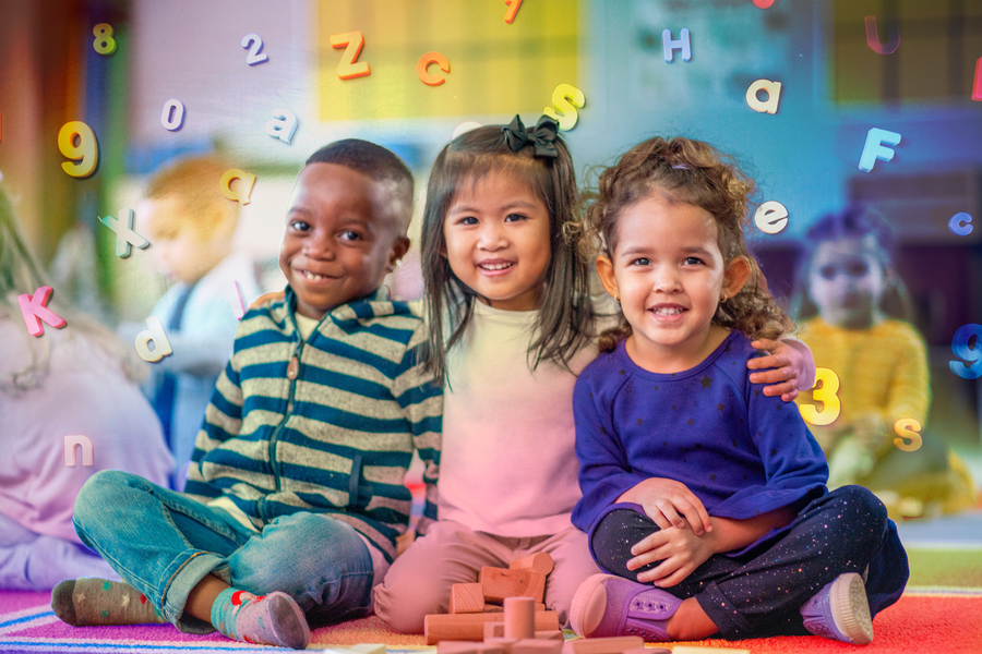 Three young children sit together on a colorful classroom floor smiling at the camera, surrounded by floating illustrated letters and numbers.